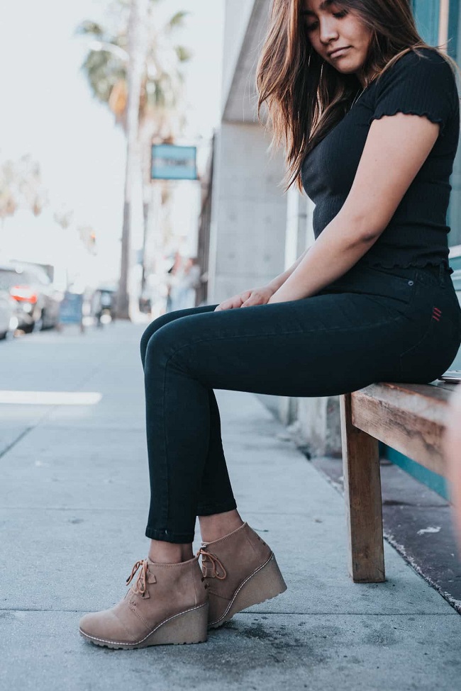 picture of a woman sitting on a wooden bench on a sidewalk wearing black pants an shirt and brown comfortable wedges
