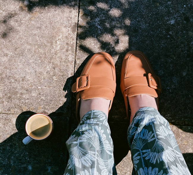 picture of a woman feet beside a cup of coffee wearing pants with flowers and orange leather  loafers 