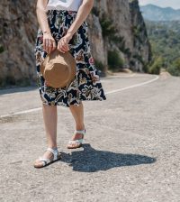 picture of a woman walking on a road in the mountain wearing skirt and flats, holding a hat